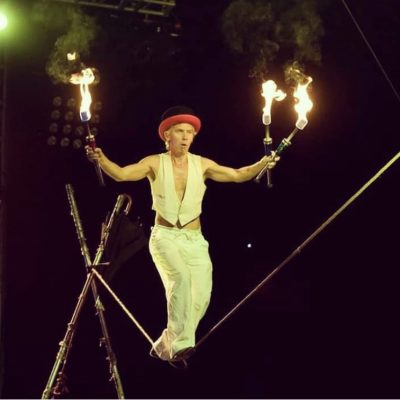 Tightrope walker on a tightrope holding up three flaming torches in the air wearing a red hat and white outfit