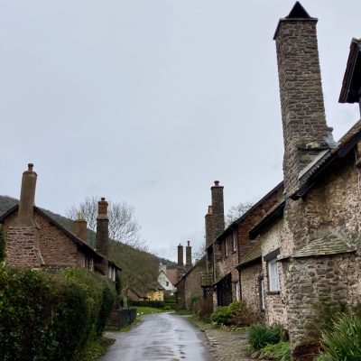 A picture of the road through Bossington with cute stone houses on either side and a yellow house in the distance and green hedges