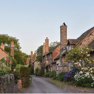 A picture of a Bossington Street with sweet colourful flowers in bloom on bushes and in flowerbeds and chimneys catching the evening sunlight