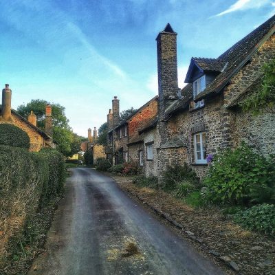 A picture of a Bossington village street with cute stone houses and trees and bushes and a blue sky in the background