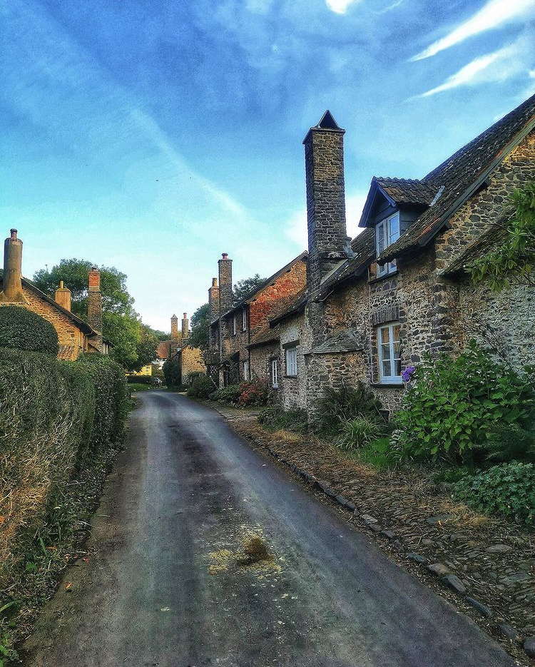 A picture of a Bossington village street with cute stone houses and trees and bushes and a blue sky in the background