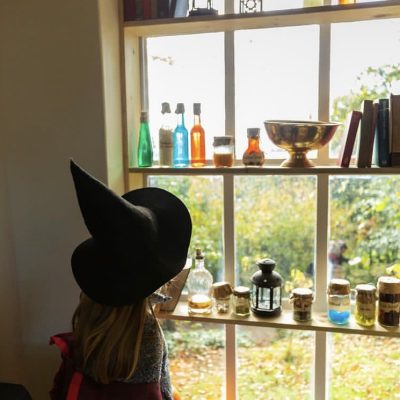 Child in witches hat at Halloween looking out of a window with bottles lining its ledges for potion making