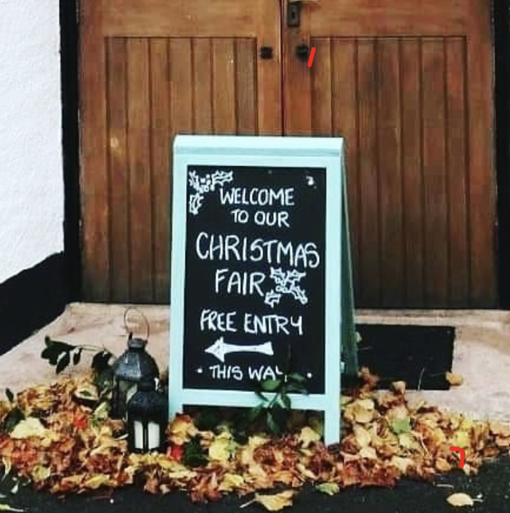 Black sandwich board in pale blue green frame with writing on it which reads "Christmas Fair Free Entry, this way" - it stands with autumn leaves at its feet and wooden doors in the background