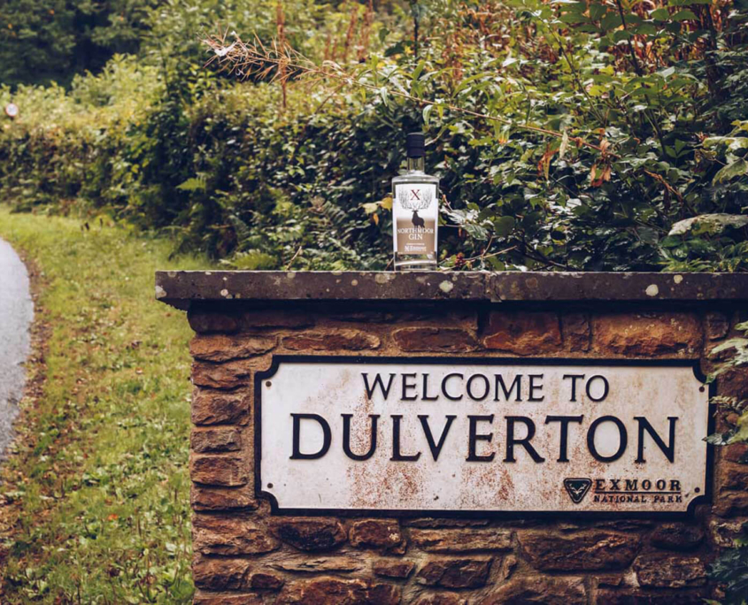 Red stone wall in countryside greenery with traditional British "Welcome to Dulverton" sign - white with black writing with a bottle of Northmoor Gin perched on the wall