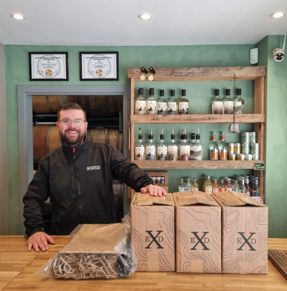 The Exmoor Distillery Shop with friendly member of staff packaging up the customer's order in brown paper bags, the wall behind him is a mint green and has wooden shelves with bottles of Exmoor Distillery booze lined up