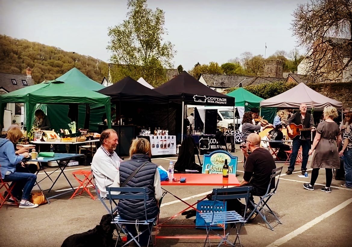 People sat at red & blue coloured tables and chairs with the black and green canvas rooves of gazebos and stands selling goods at Dulverton Farmers Market