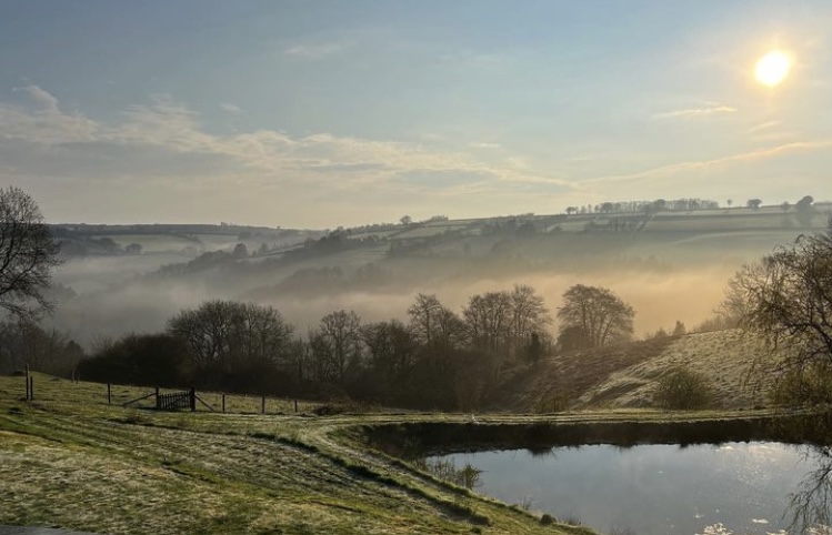 Landscape of the rolling hills of Exmoor, near Dulverton with a lake in the foreground and the sun shining through a misty sky