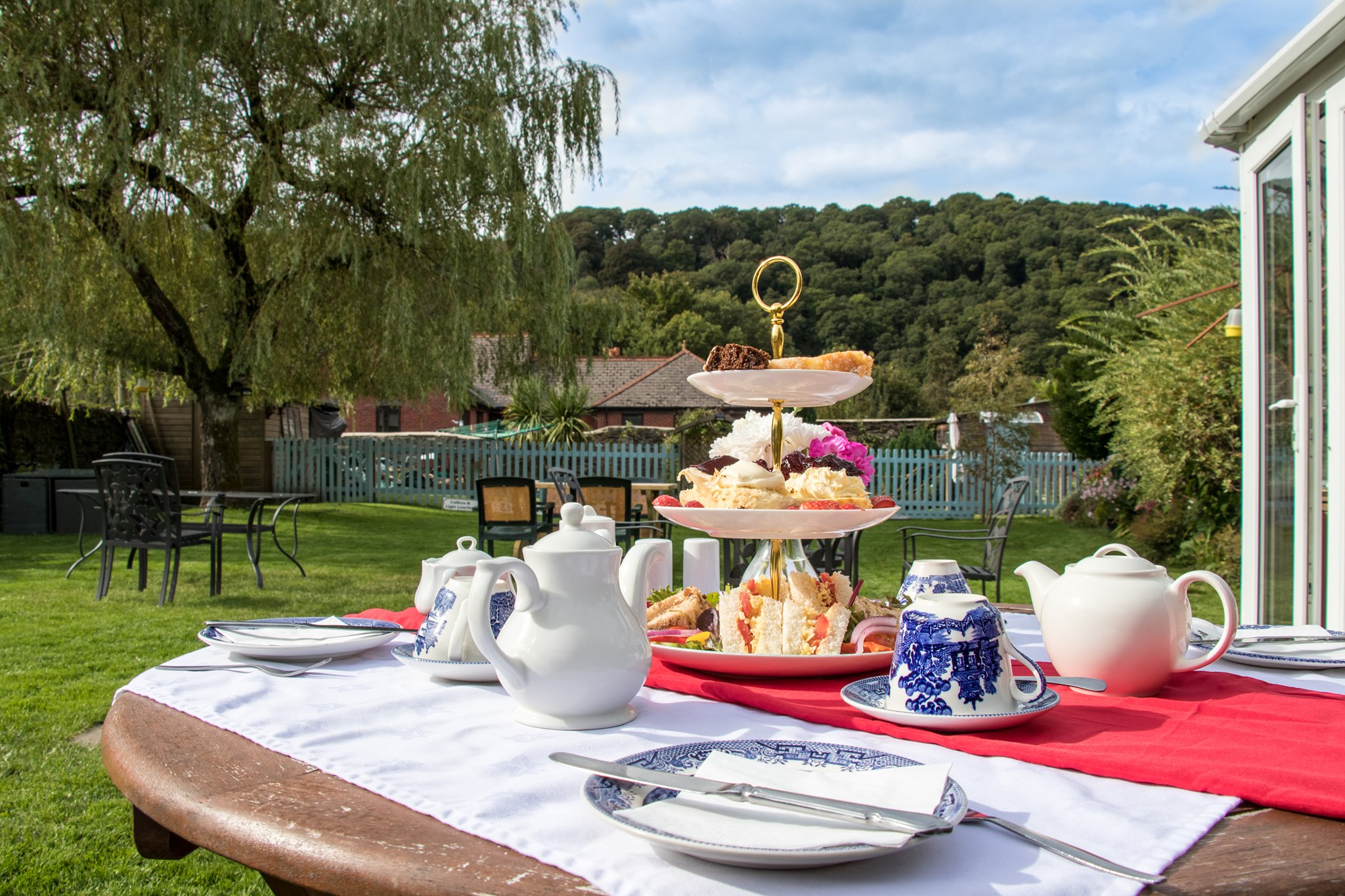 The Copper Kettle Cream Tea Outside in the garden with sandwiches, scones & a view of a pretty garden behind the table with cute china pattern crockery
