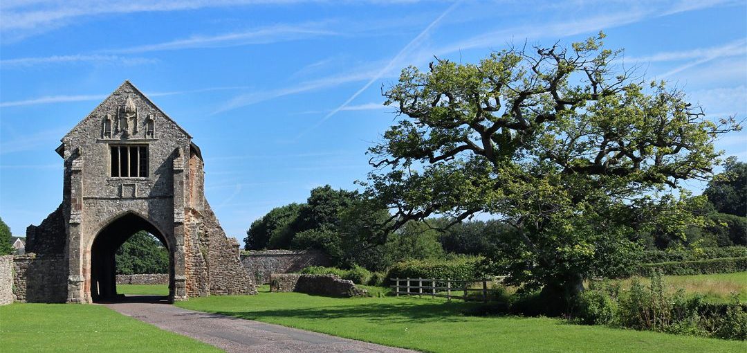 The entrance to Cleeve Abbey on a sunny day, blue skies with streaky clouds in the background, lush green grass in the foreground, trees to the right and the arched ancient stone door way to the left