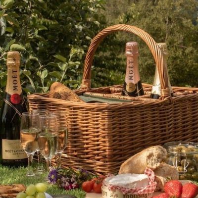 Wicker picnic hamper with bottles of Moet Champagne and a picnic laid out in front of it, greenery behind it