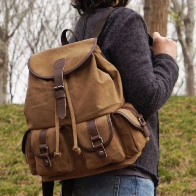 Brown canvas buckle up backpack slung over a woman's shoulder - we see her midsection with a green field and trees behind her