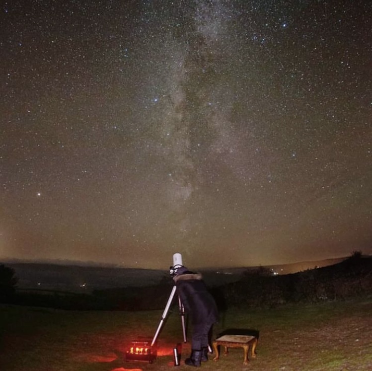 A stargazer looking through a telescope in Exmoor National Park - glowing red lights around the telescope and a sky filled with thousands of stars