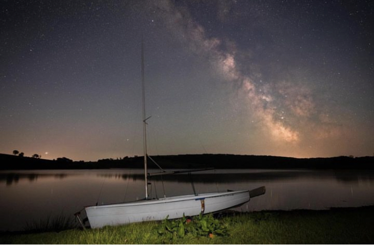 Stargazing on Exmoor at Wimbleball Lake with white wooden boat in the foreground on the bank of the lake, and the incredible milkyway and night sky reflecting in the surface of the lake