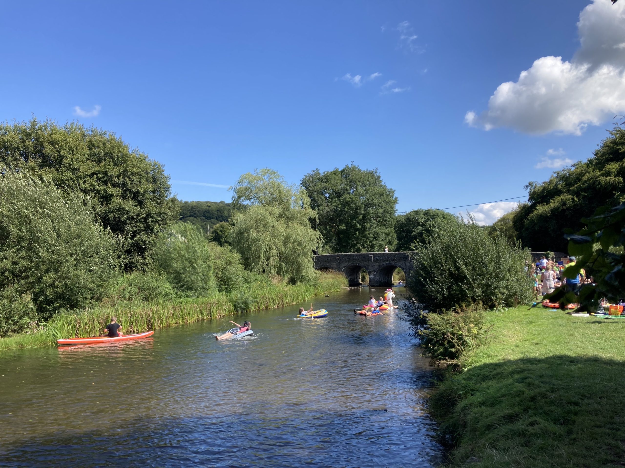 People swimming & canoeing on the river near Withypool in Exmoor, arched bridge in the distance, sunny weather shining on the river and grassy banks