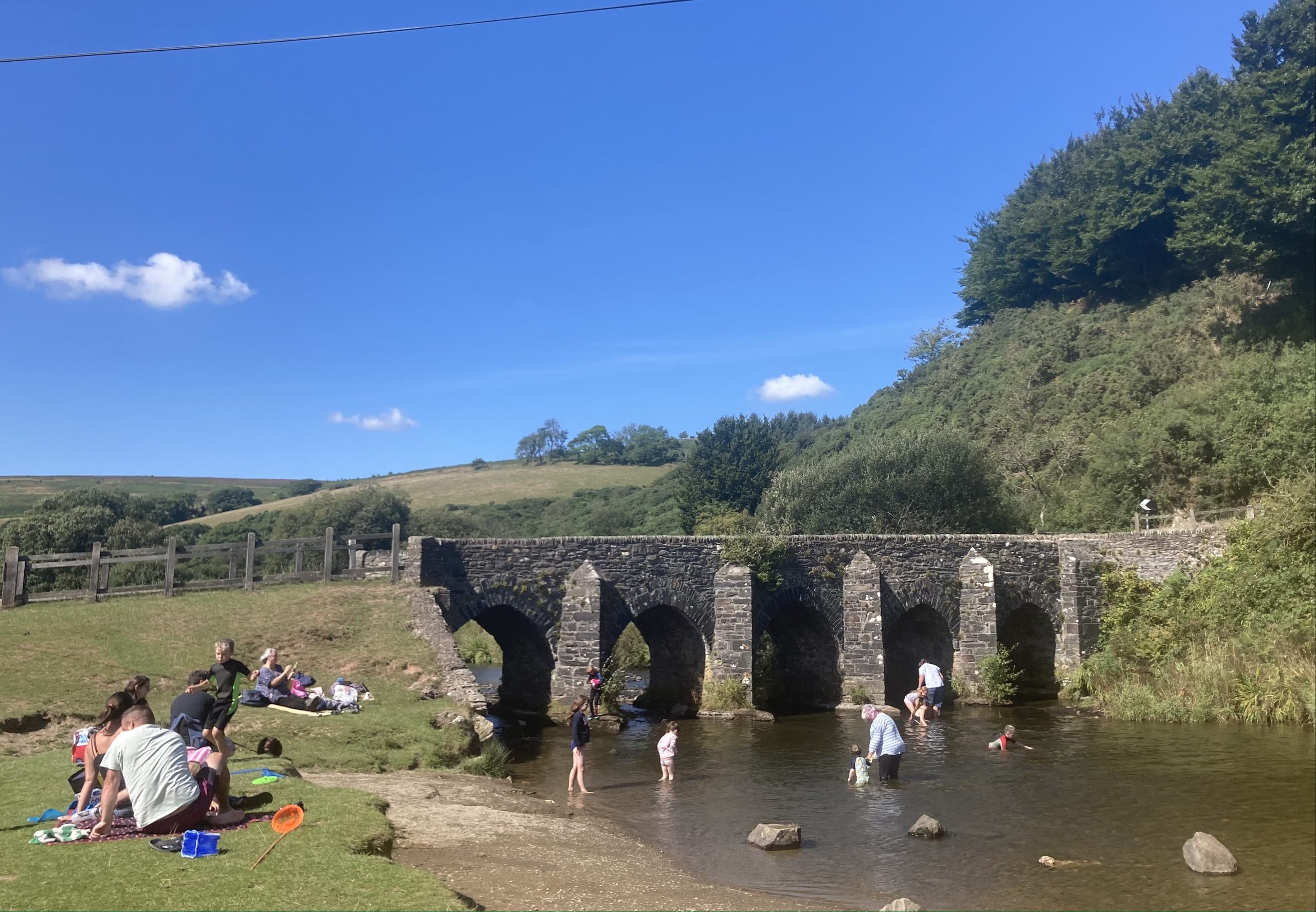 People Wild Swimming at Landacre Bridge near Withpool, a beautiful old arched bridge with clear water river flowing through it and grassy banks - blue skies and green countryside