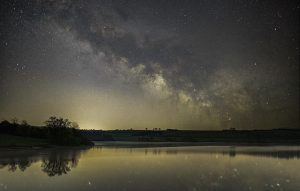 Stargazing on Exmoor - dark blue night sky with yellow golden glimmers dotted with stars, a silhouetted countryside landscape and a lake reflecting the sky